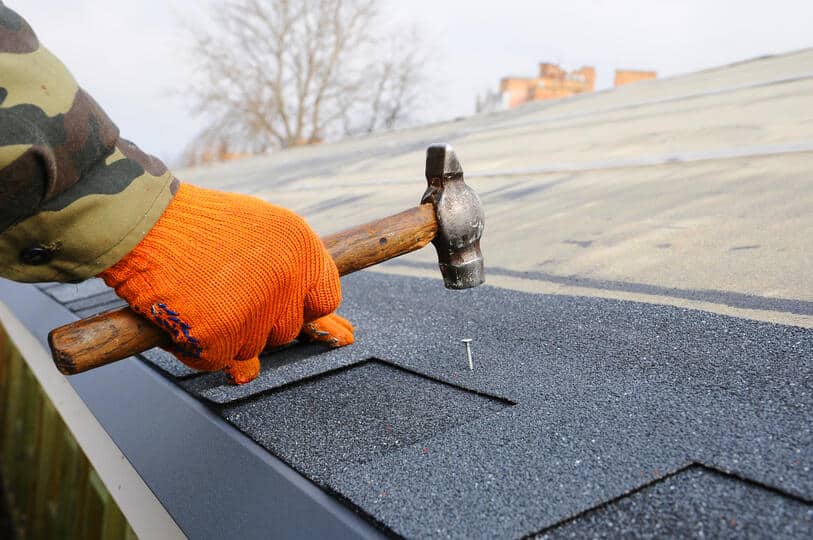 how to start shingles on a roof-close up of a roof tech wearing gloves using a hammer to nail a strip of shingles to a roof