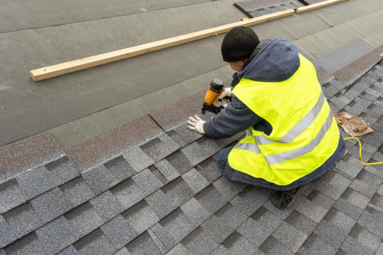 how to start shingles on a roof-over the shoulder view of a roof tech in a safety vest using a power tool to nail shingles to roof